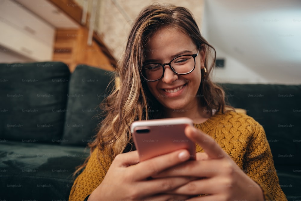 Young woman using smartphone and smiling while sitting on the floor at home and wearing eyeglasses