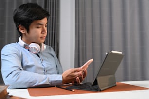 Casual asian man with headphone sitting in front of his computer tablet and using smart phone.