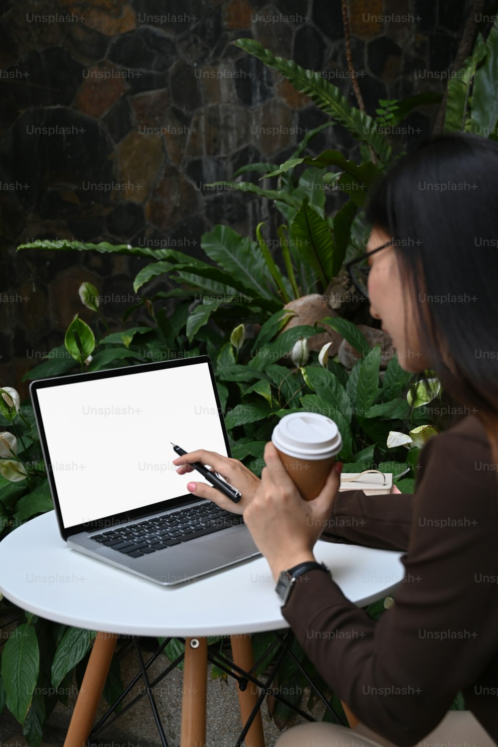 Side view of businesswoman in glasses holding cup of coffee and working with laptop computer at cafe.