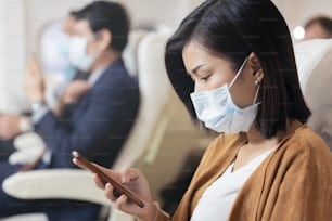 Passenger wearing face mask using mobile phone on airplane during covid pandemic to prevent coronavirus infection