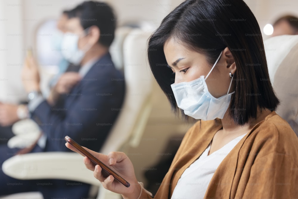 Pasajero con mascarilla usando teléfono móvil en el avión durante la pandemia de covid para prevenir la infección por coronavirus