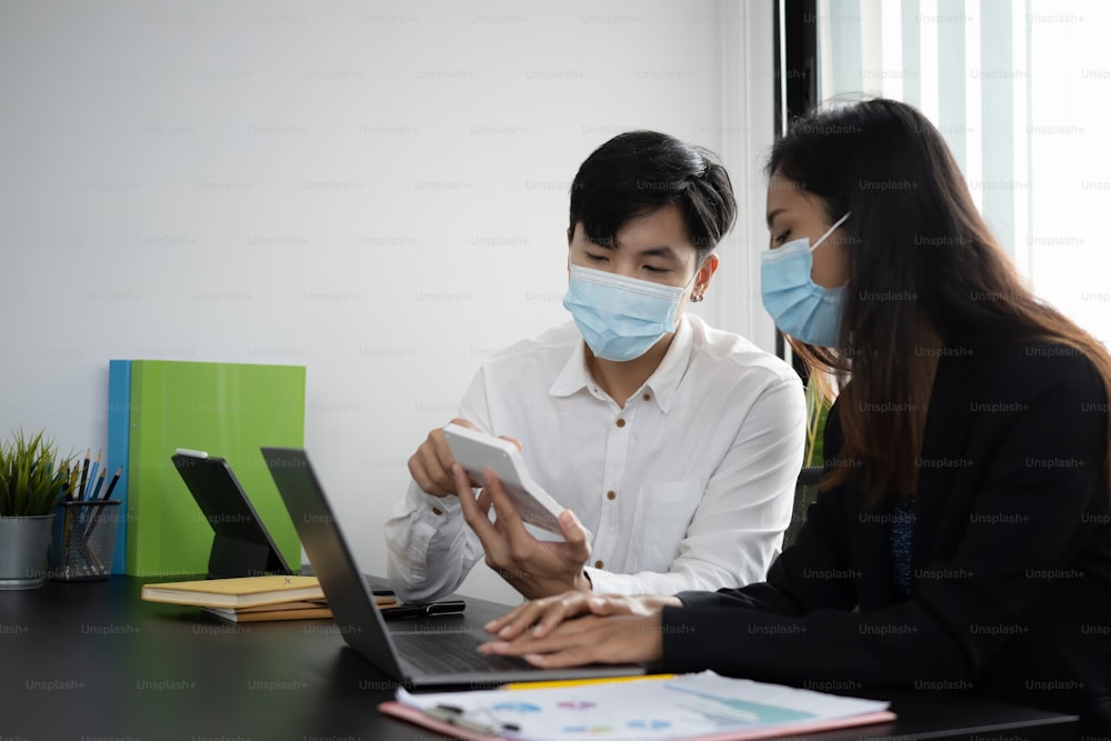 두 사업가는 보호 마스크를 쓰고 계산기를 사용하여 사무실에서 함께 일하고 있습니다.