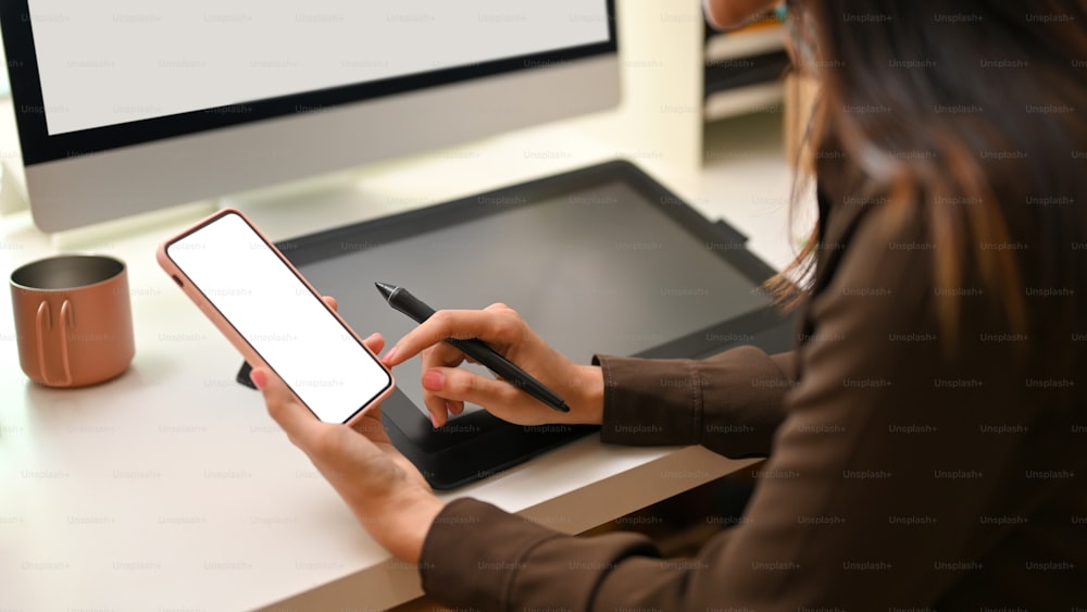 Beschnittene Aufnahme einer Designerin mit dem Smartphone während der Arbeit am Büroschreibtisch, Clipping-Pfad