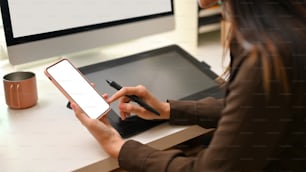 Beschnittene Aufnahme einer Designerin mit dem Smartphone während der Arbeit am Büroschreibtisch, Clipping-Pfad