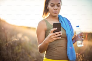 Sporty young woman holding bottle of water and using mobile. Focus is on hand.