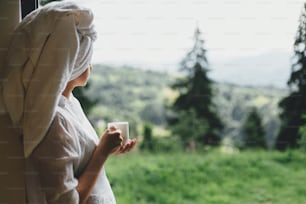 Beautiful young woman enjoying sunny morning with cup of coffee on background of mountain hills. Tranquil moment, space for text. Beautiful relaxed female in white pajamas and towel drinking coffee