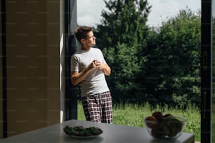 Handsome man enjoying sunny morning with cup of coffee on porch on background of mountain hills. Tranquil moment. Space for text. Young man in pajamas drinking coffee and relaxing
