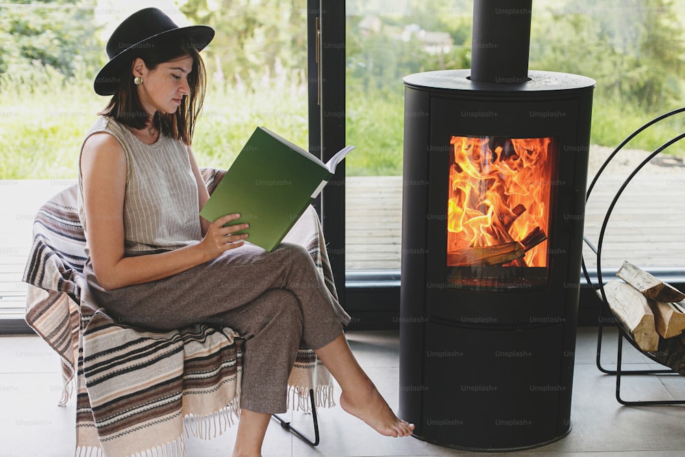 Beautiful stylish woman reading book on chair at warm burning fireplace on background of big window. Young female in casual clothes relaxing in modern chalet, cozy vacation. Leisure time