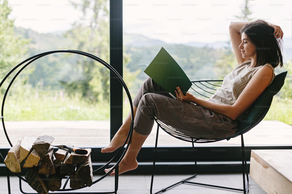 Beautiful stylish woman reading book on chair at firewood on background of big window with view on mountain hills. Young female in casual clothes relaxing in modern chalet. Leisure time