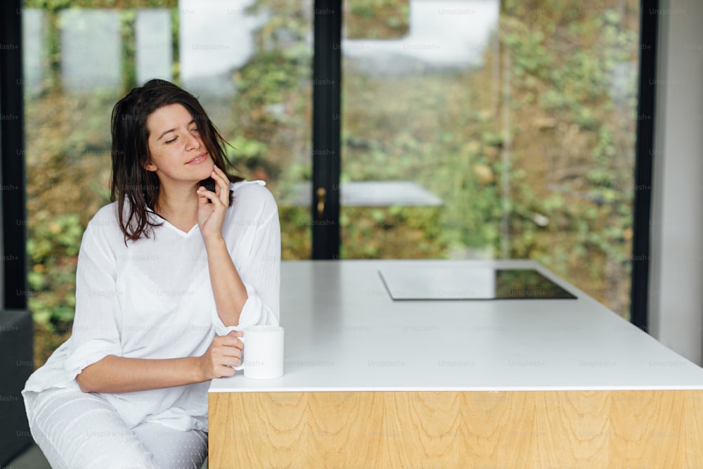 Beautiful happy woman enjoying hot coffee in sunny morning  in modern kitchen in chalet in mountains. Calm carefree moment. Young female in white pajamas relaxing with cup of coffee