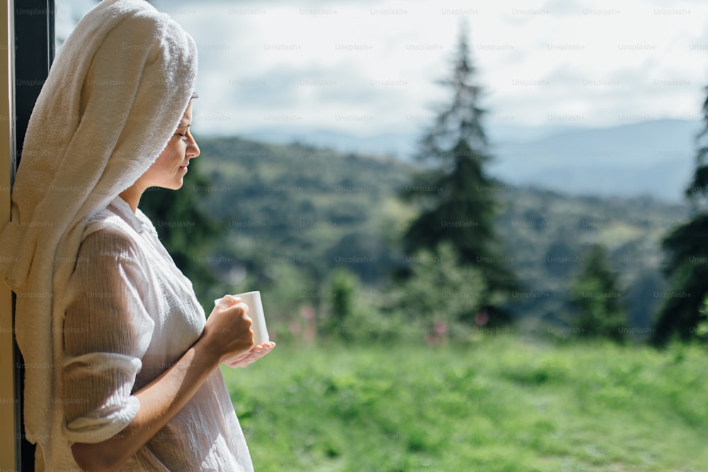 Beautiful young woman enjoying coffee in sunny morning on background of mountain hills. Calm tranquil moment. Beautiful female in white pajamas and towel after shower relaxing with cup of coffee