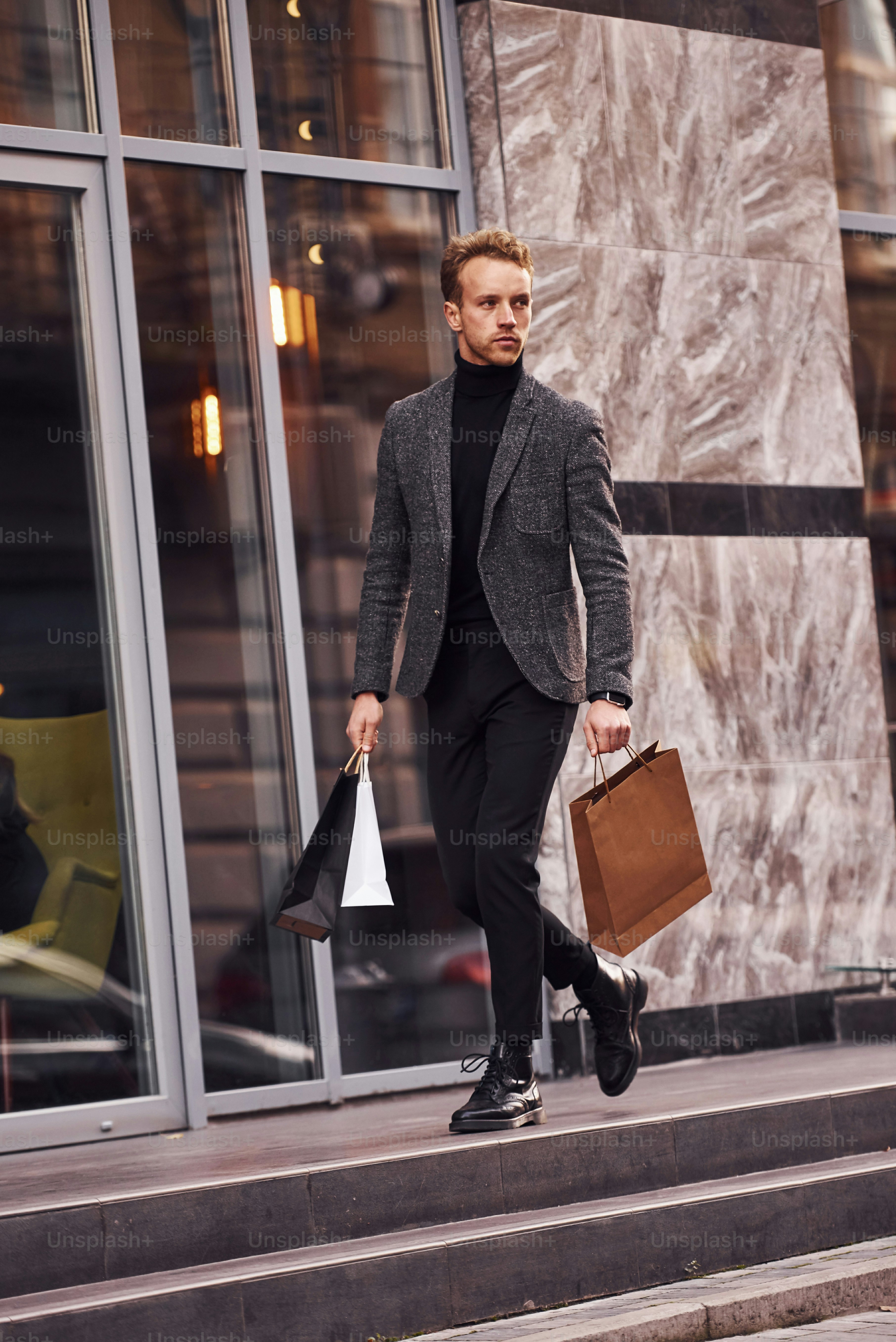 Man in elegant formal wear and with shopping packages is outside against modern building.