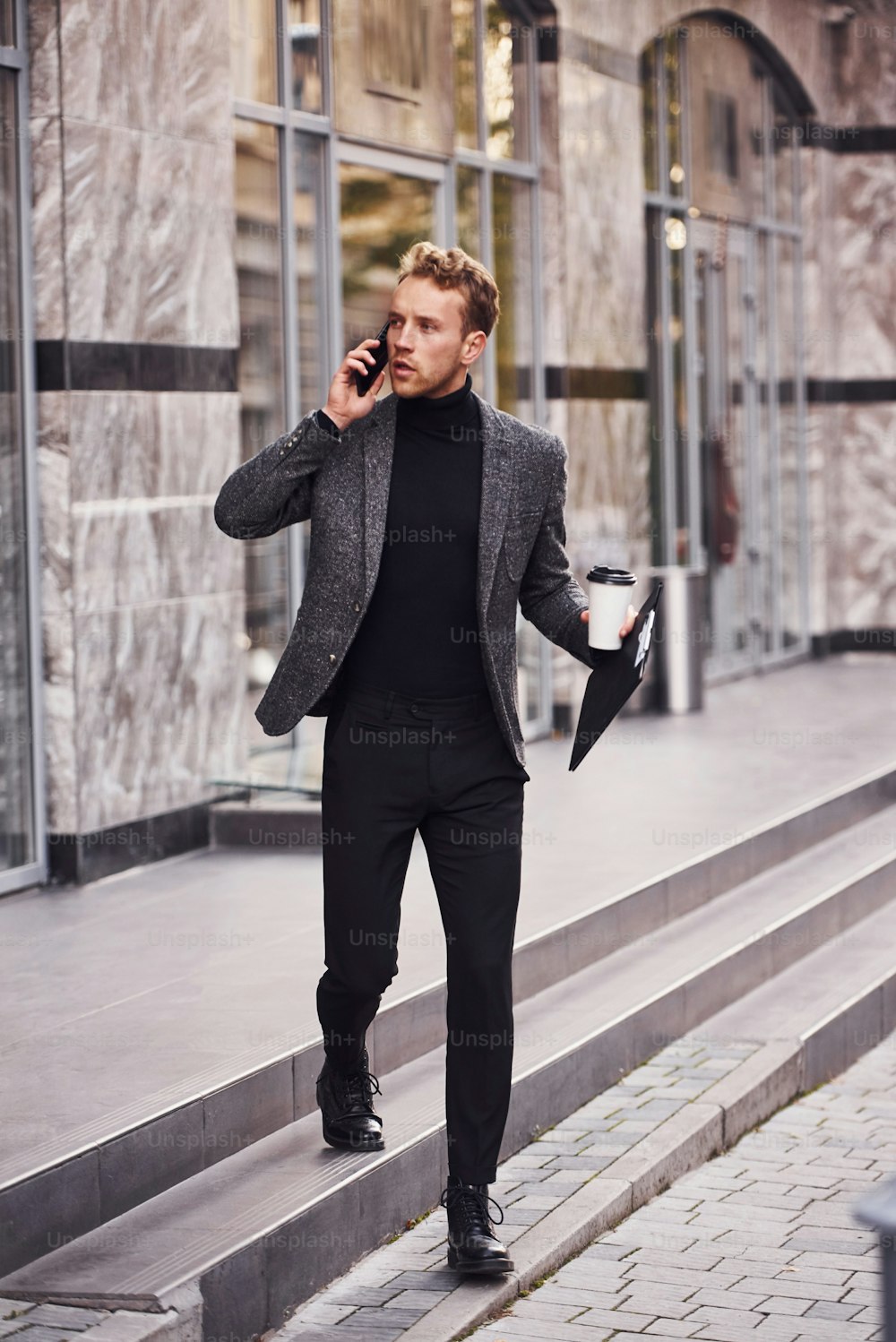 Man in elegant formal wear with cup of drink and notepad in hands is outside against modern building have a conversation by the phone.