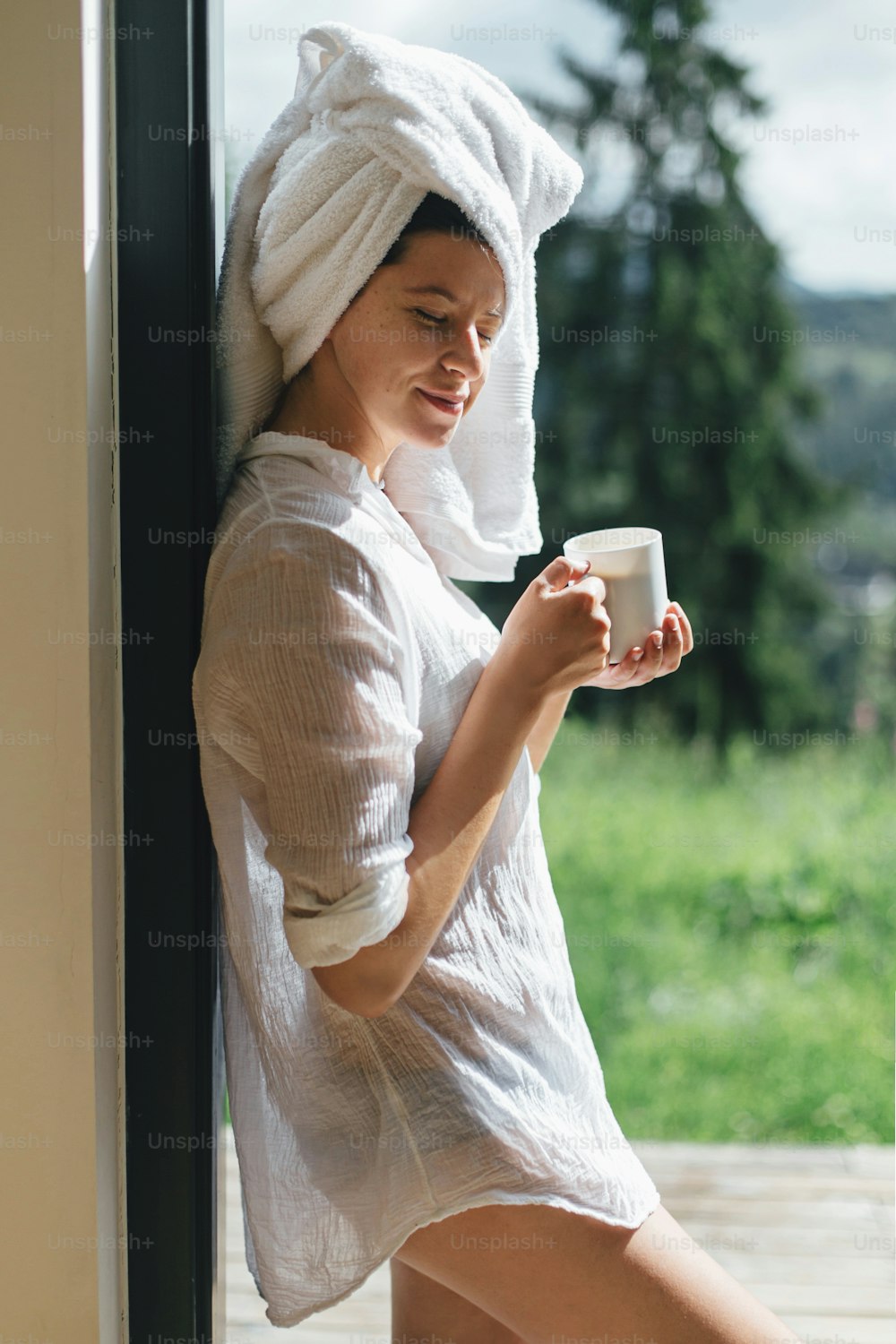 Beautiful young woman enjoying sunny morning with cup of coffee on background of mountain hills. Tranquil moment, space for text. Beautiful relaxed female in white pajamas and towel drinking coffee