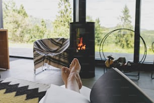 Beautiful female feet and book on background of warm burning fireplace and big window in modern chalet. Young woman barefoot relaxing with book in comfortable home, cozy vacation. Leisure time