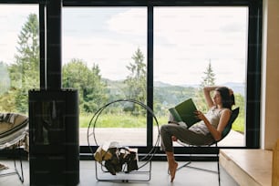 Beautiful stylish woman reading book on chair at fireplace with firewood on background of mountain hills. Young female in casual clothes relaxing in modern chalet with amazing view from window