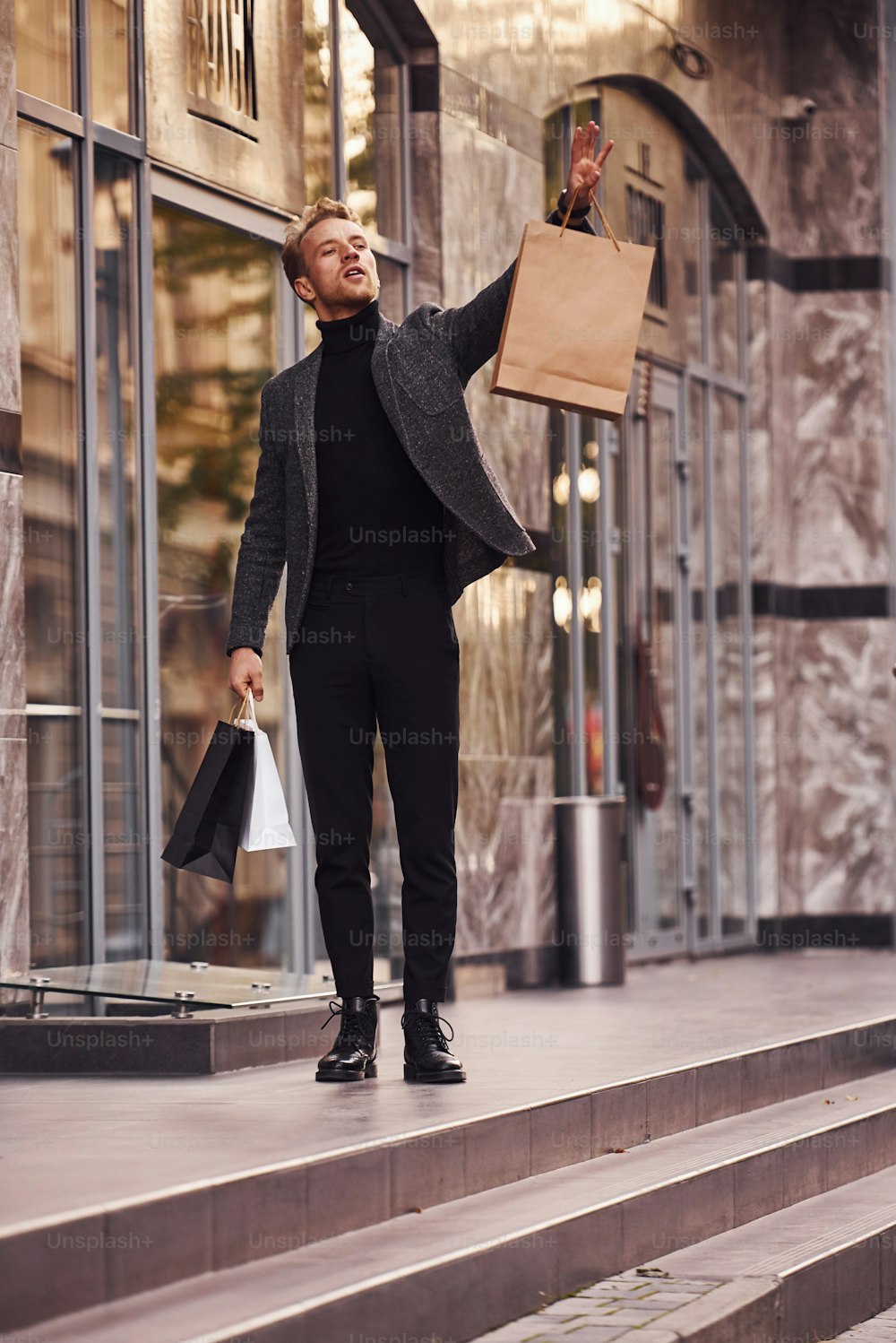 Homem em trajes formais elegantes e com pacotes de compras está do lado de fora contra a construção moderna.