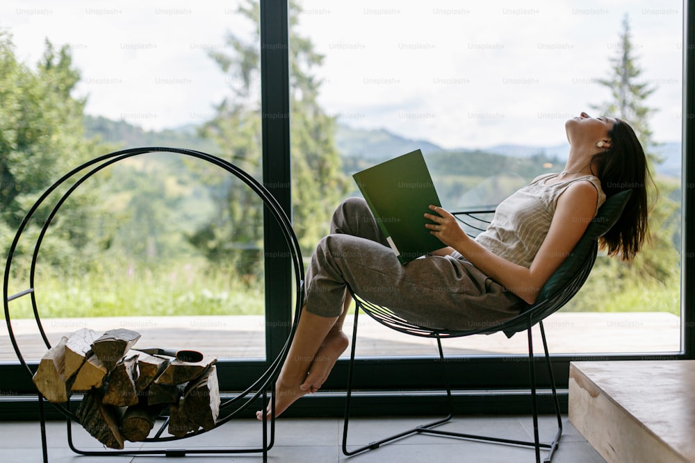Beautiful stylish woman relaxing with book in modern chalet with amazing view on mountain hills. Leisure time. Young female in casual clothes reading book on chair at firewood.