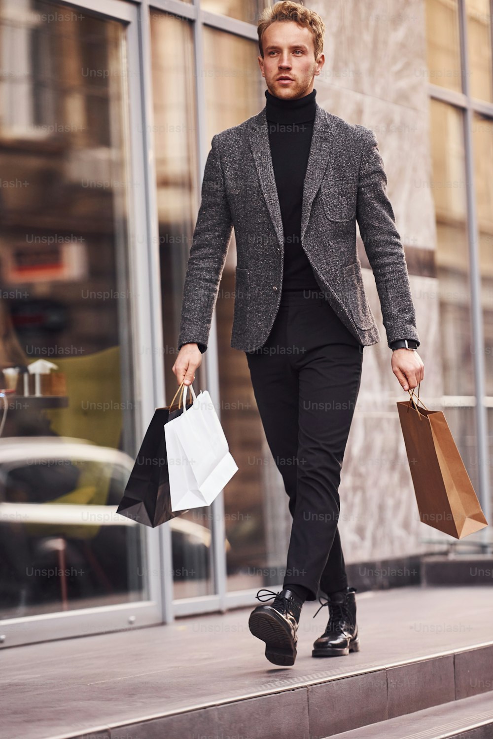 エレガントなフォーマルウェアを着て、ショッピングパッケージを持った男性が、近代的な建物を背景に外にいます。