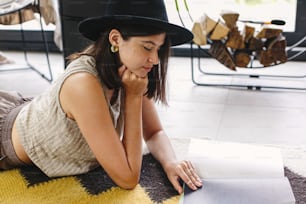 Beautiful stylish woman reading book on floor on background of fireplace and big window in modern chalet. Young female in casual clothes relaxing on cozy rug with magazine. Leisure and vacation time