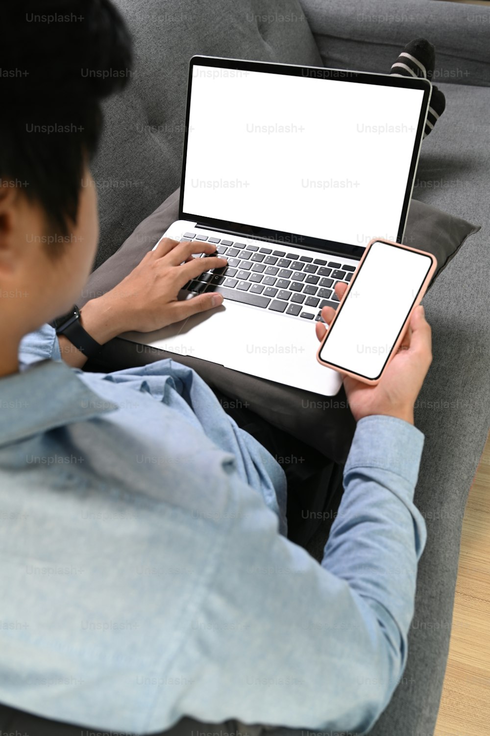 Vista sobre el hombro de un joven que descansa en el sofá que trabaja con una computadora portátil y usa un teléfono móvil.