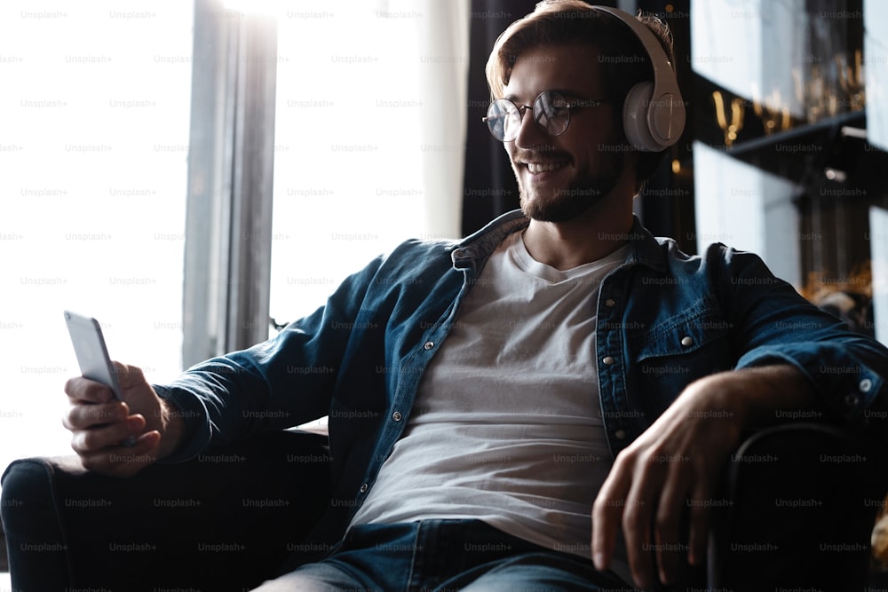 Smiling young adult man wearing wireless headphones listening mobile music on smartphone, happy guy using online player podcast audio book in app on cell phone relaxing at home