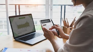 Young woman accountant using calculator and analyzing company annual financial report at office desk.