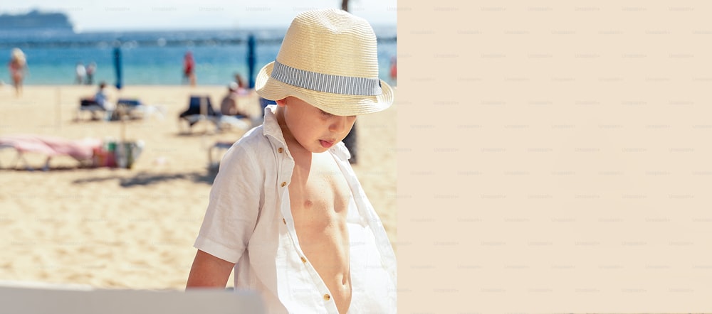 Garotinho elegante no chapéu brincando na praia tropical arenosa. Vibrações de verão. Muito espaço de cópia.