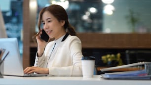 Side view of young businesswoman talking on the phone while working with laptop in modern office room