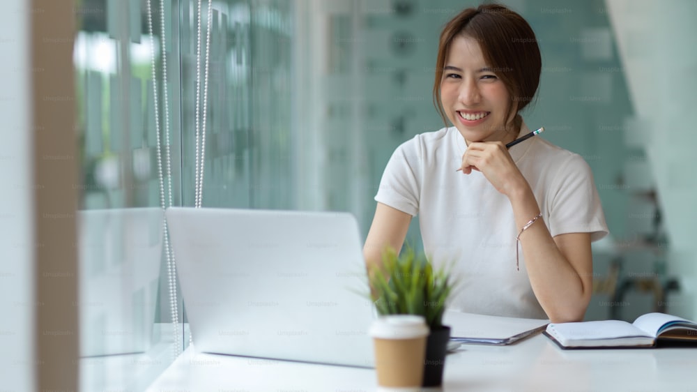 Half-length portrait of friendly young pretty female laughing and looking into camera while working in modern workplace