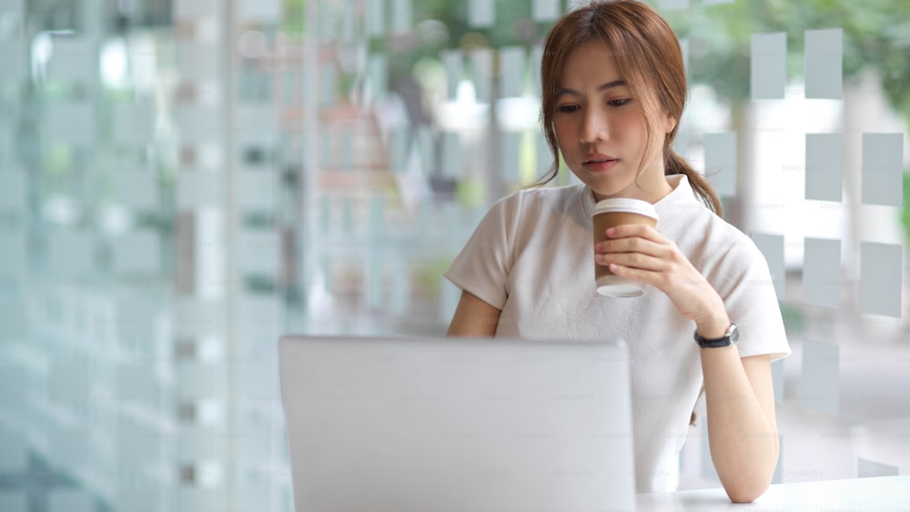 Halbfigurenporträt einer jungen, hübschen Frau, die eine Kaffeetasse hält und im Coworking Space mit einem Laptop arbeitet