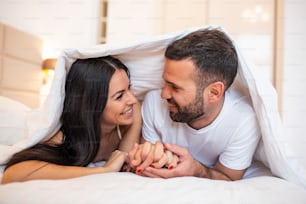 smiling couple going to kiss under sleeping sheets in the morning. In love young happy couple lying in bed in the morning