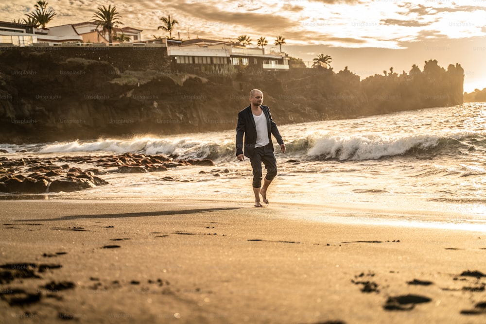 Young businessman walking on the sandy beach during beautiful golden sunset, wearing suit.