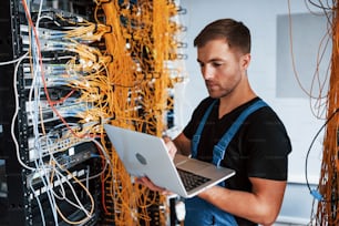 Young man in uniform and with laptop works with internet equipment and wires in server room.