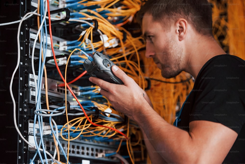 Young man in uniform works with internet equipment and wires in server room.