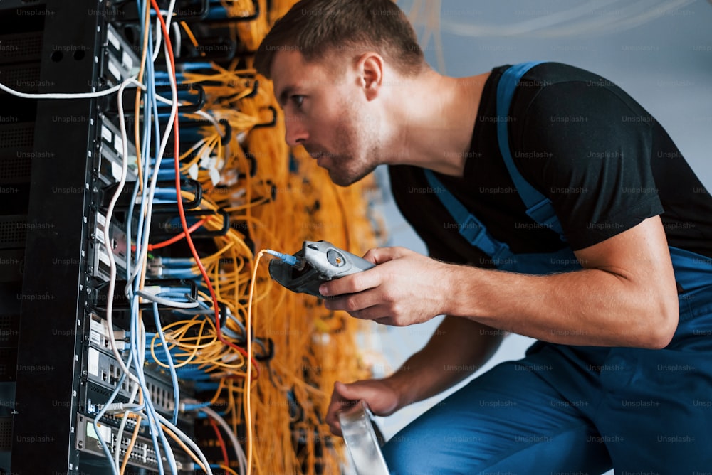 Young man in uniform with measuring device works with internet equipment and wires in server room.