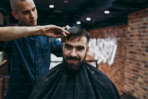 Hombre adulto joven guapo que recibe un tratamiento de peinado y vestimenta para el cabello y la barba por parte de un estilista profesional.