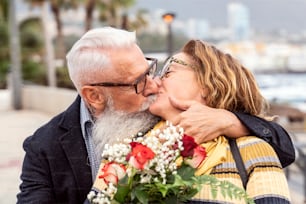 Portrait of lovely attractive cheerful couple of seniors hugging and kissing during the anniversary date. Handsome man surprised his wife with flowers on valentine's day. Real people emotions.