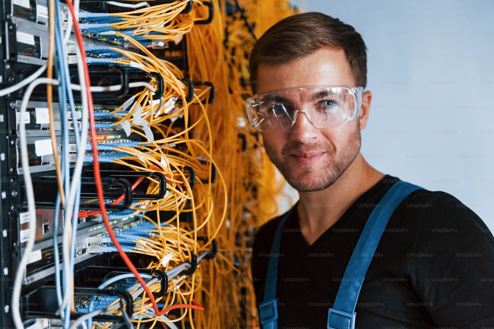 Young man in protective glasses works with internet equipment and wires in server room.