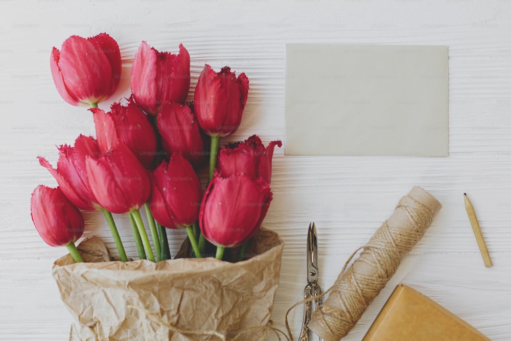 Beautiful red tulips in craft paper with greeting card, gift, pencil and scissors on white wooden background. Rustic flat lay, Greeting card template. Happy Mothers day. Happy Women's day.