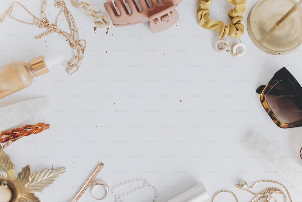 Modern summer accessories flat lay with space for text. Feminine essentials. Gold jewellery, sunglasses, hair clips, barrettes, cosmetics on white table with vintage candles