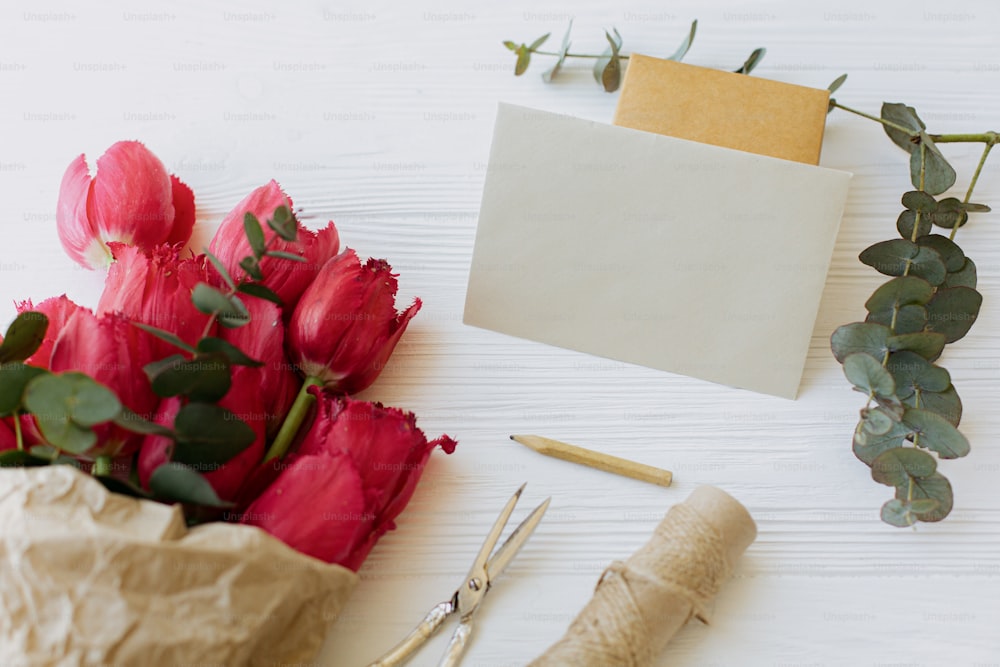 Beautiful red tulips in craft paper with greeting card, gift, pencil, scissors and twine on rustic white wooden background. Greeting card template. Happy Mothers day. Happy Women's day.