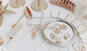 Modern summer accessories. Golden jewellery on boho mirror, barrettes, hair clips, cosmetics, perfume and lace lingerie on white table with vintage candles. Feminine essentials