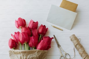 Preciosos tulipanes rojos en papel artesanal con tarjeta de felicitación, regalo, lápiz, tijeras y cordel sobre fondo rústico de madera blanca. Plantilla de tarjeta de felicitación. Feliz día de la madre. Feliz día de las mujeres.