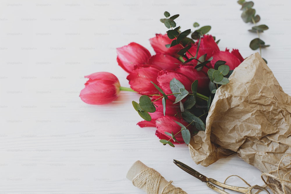Beau bouquet de tulipes rouges et d’eucalyptus en papier kraft avec des ciseaux et de la ficelle sur fond en bois blanc rustique. Modèle de carte de voeux. Joyeuse Journée de la Femme. Bonne fête des mères