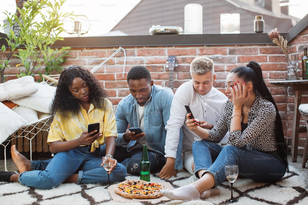 Vier multirassische Menschen in stilvoller Kleidung sitzen zusammen auf dem Dach und benutzen persönliche Smartphones. Junge Freunde trinken Alkohol und essen Pizza. Moderner Lebensstil.