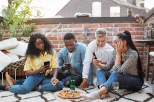 Four multiracial people in stylish clothes sitting together on rooftop and using personal smartphones. Young friends drinking alcohol and eating pizza. Modern lifestyle.