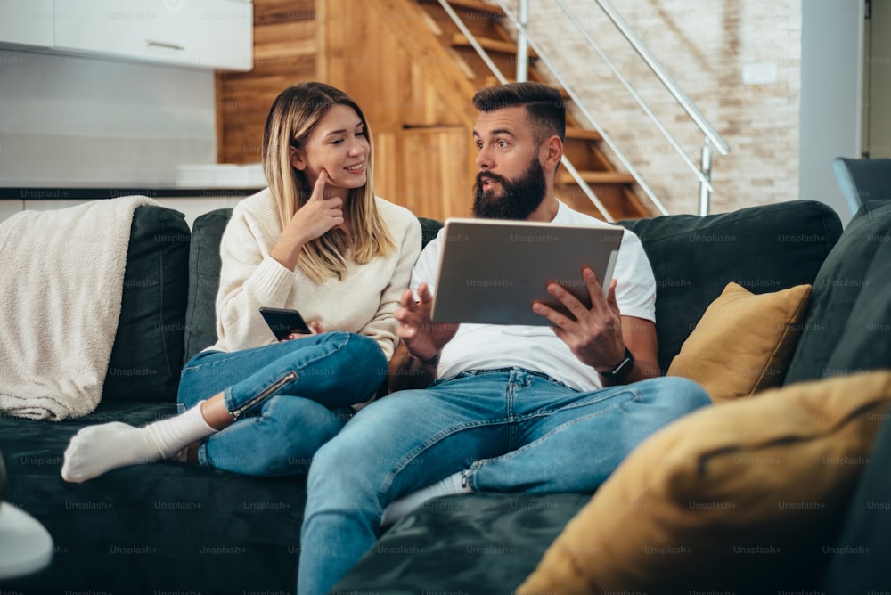 Jovem casal atraente usando um smartphone e um laptop enquanto relaxa no sofá em casa