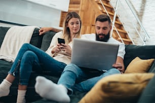 Young couple suing a smartphone and a laptop while relaxing on the sofa at home