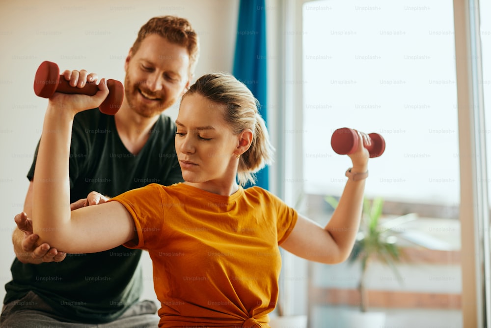 Athletic woman using dumbbells while working out with husband's assistance at home.